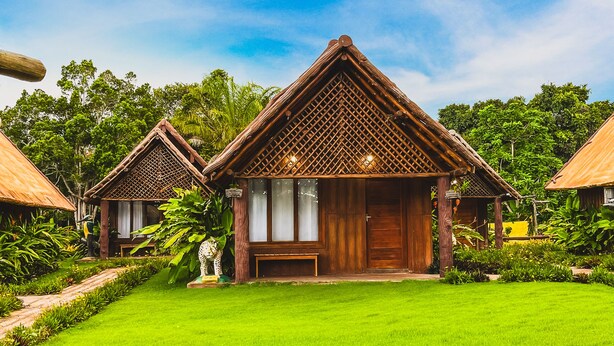 A bungalow-style resort with a thatched roof and wooden walls, surrounded by lush greenery and a well-maintained lawn. - Hotel Chalés do Santuario  (Santa Cruz Cabralia)