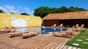 A pool area with lounge chairs, a wooden deck, and a building with a yellow wall and a white structure painted on it. - Hotel Chalés do Santuario  (Santa Cruz Cabralia)