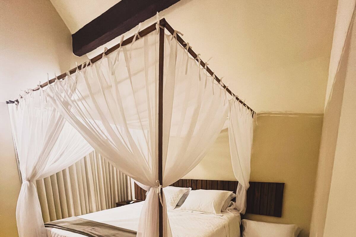 A four-poster canopy bed with a wooden bench and a small step leading to it, in a room with a sloped ceiling and a plain wall.