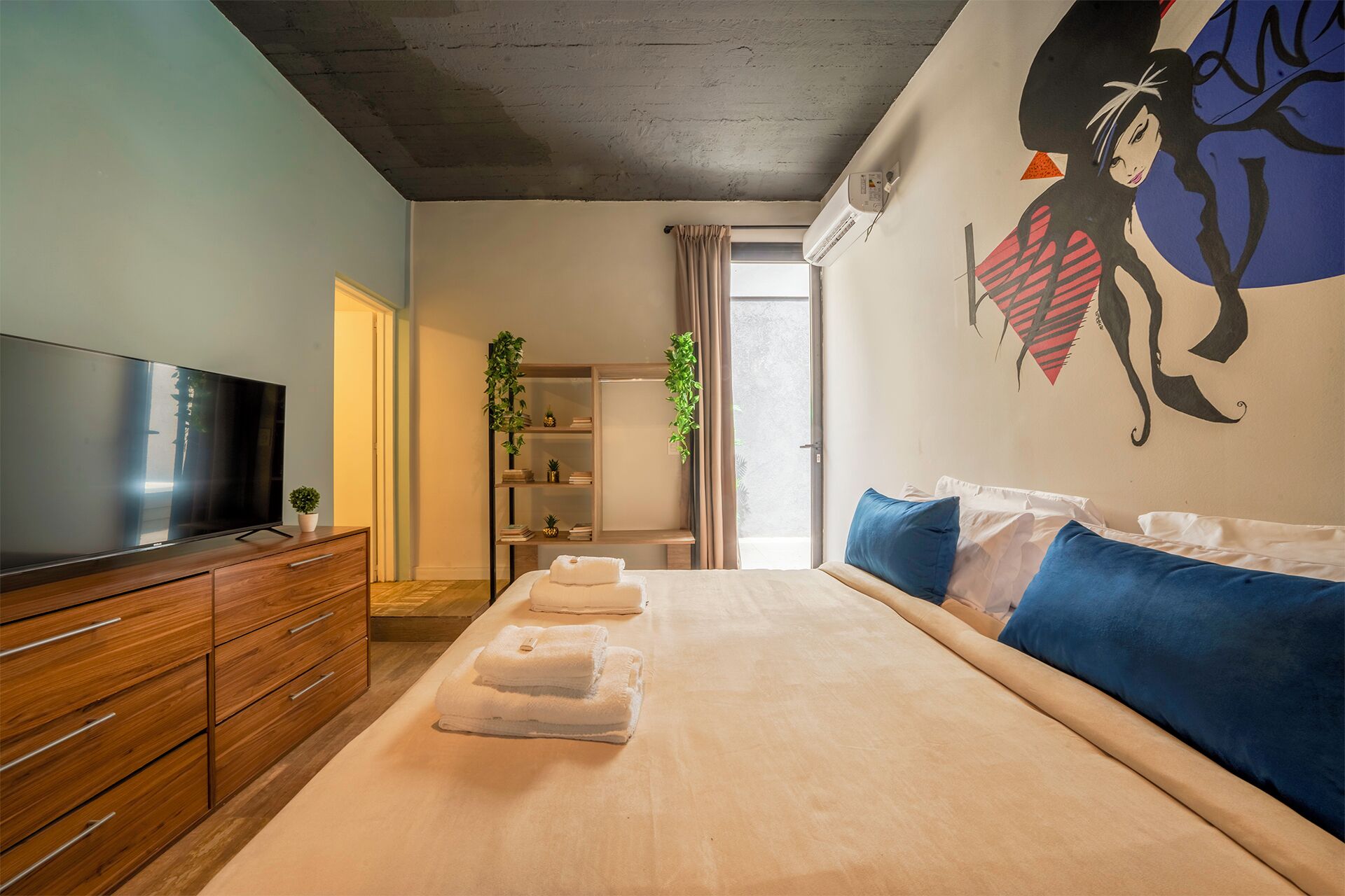 Una habitación de hotel moderna con una cama grande, un televisor de pantalla plana, muebles de madera, una planta y un mural en la pared.