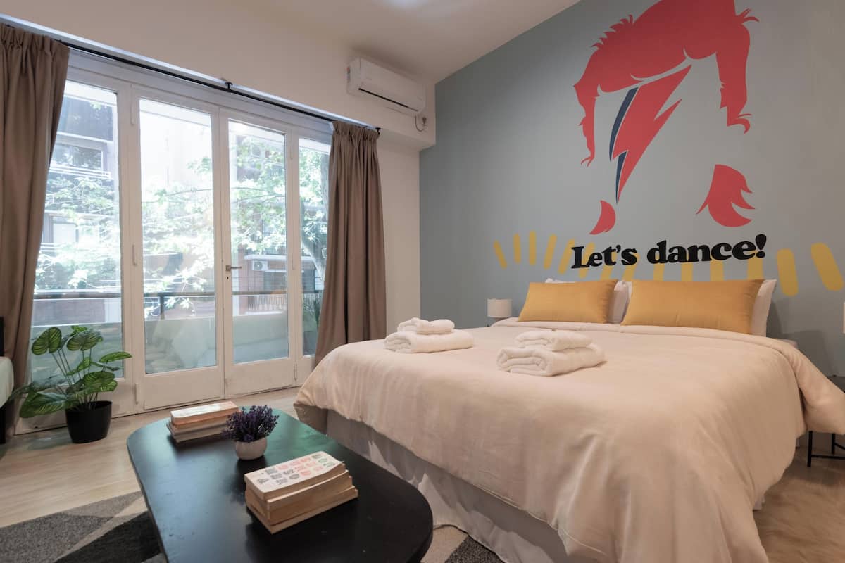 A hotel room with a bed, a small table with books and a plant, a sliding glass door to the outside, and a wall with a dance-themed mural.