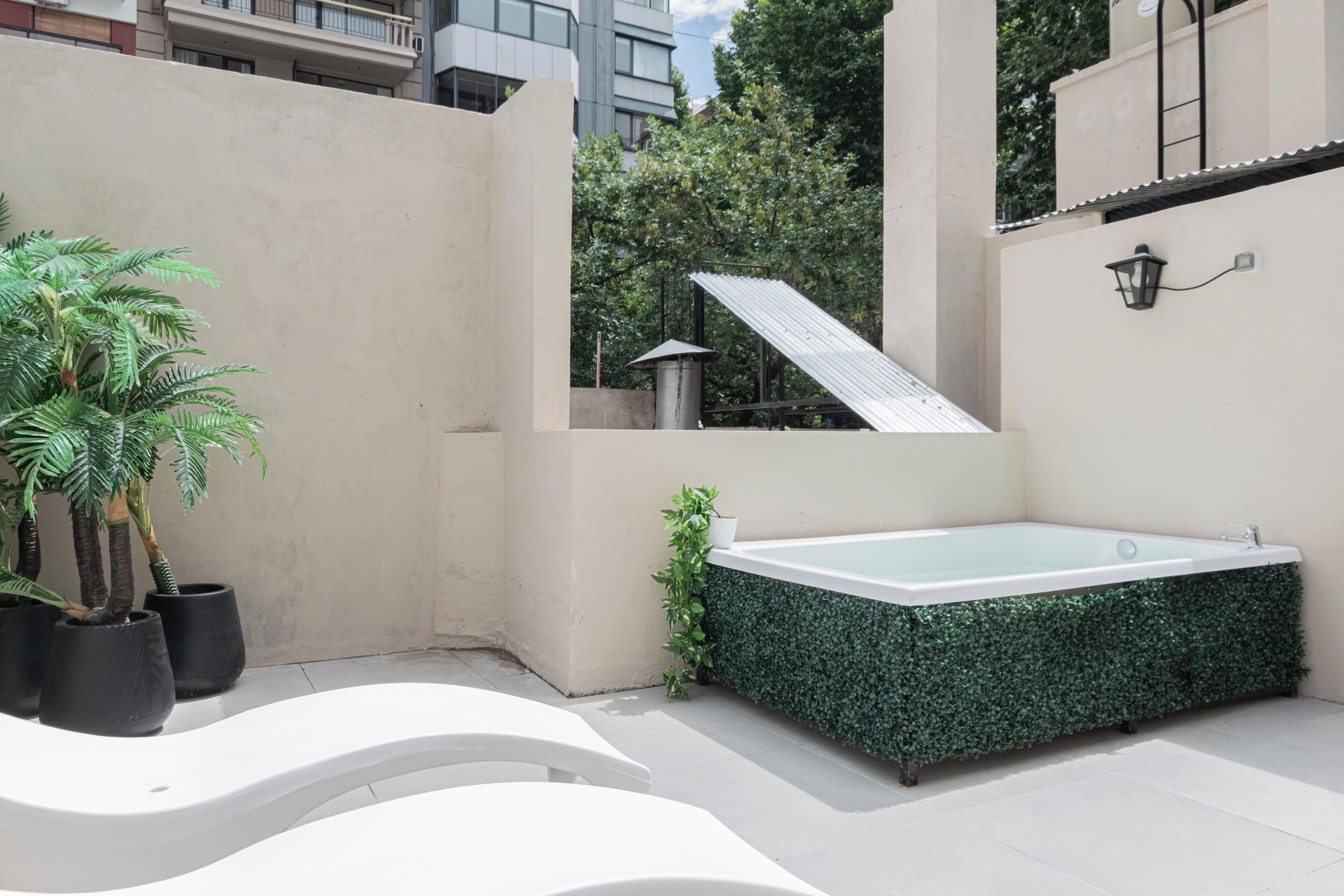 Une salle de bain extérieure moderne avec un jacuzzi rectangulaire, entouré de plantes en pot et agrémenté d’un banc blanc.