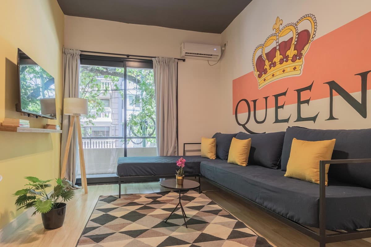 A modern living room with a sofa, a coffee table, and a potted plant. A large wall mural with a crown and the word "QUEEN" is visible.