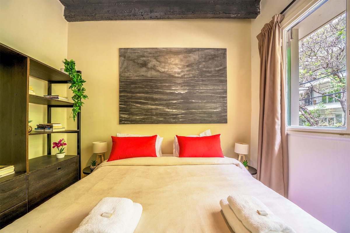 A hotel room with a bed, two red pillows, a beige blanket, a large abstract painting, a wooden wardrobe, and a window with curtains.