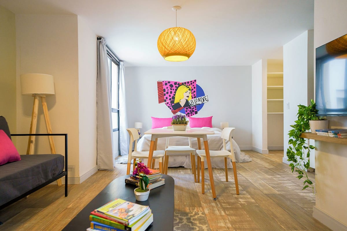 A modern living room with a sofa, coffee table, and dining area. There is a large window with curtains, a hanging light fixture, and a wall-mounted artwork.
