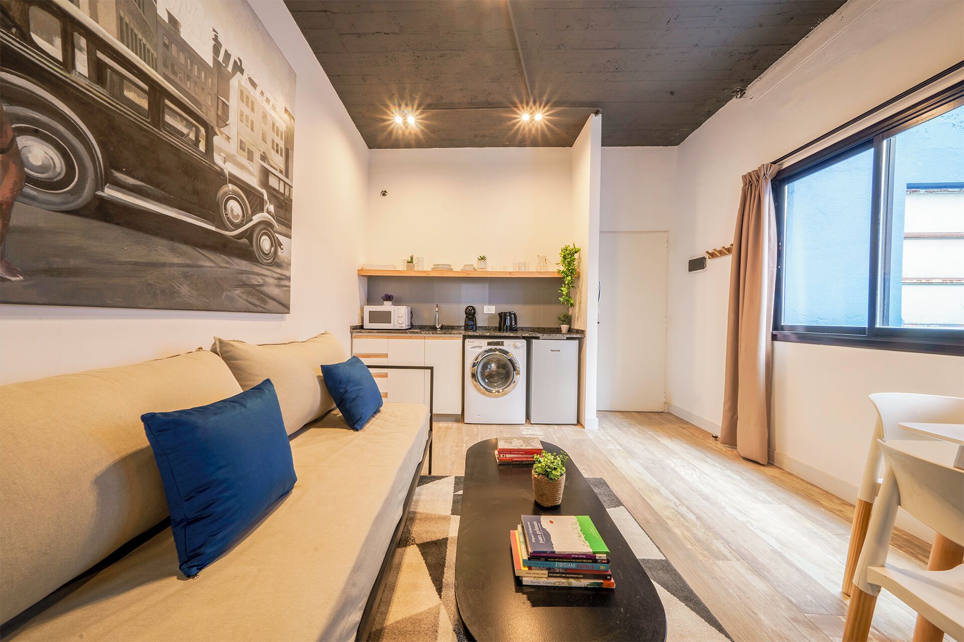 Un espacio habitable pequeño con sofá, una mesa de centro, estantería, lavadora y ventana con cortinas.