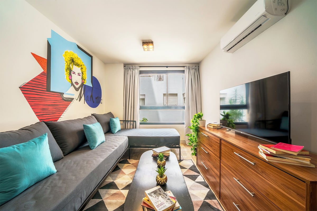 Un salon moderne avec un canapé, une table basse, un téléviseur et un mur orné d’une fresque colorée.