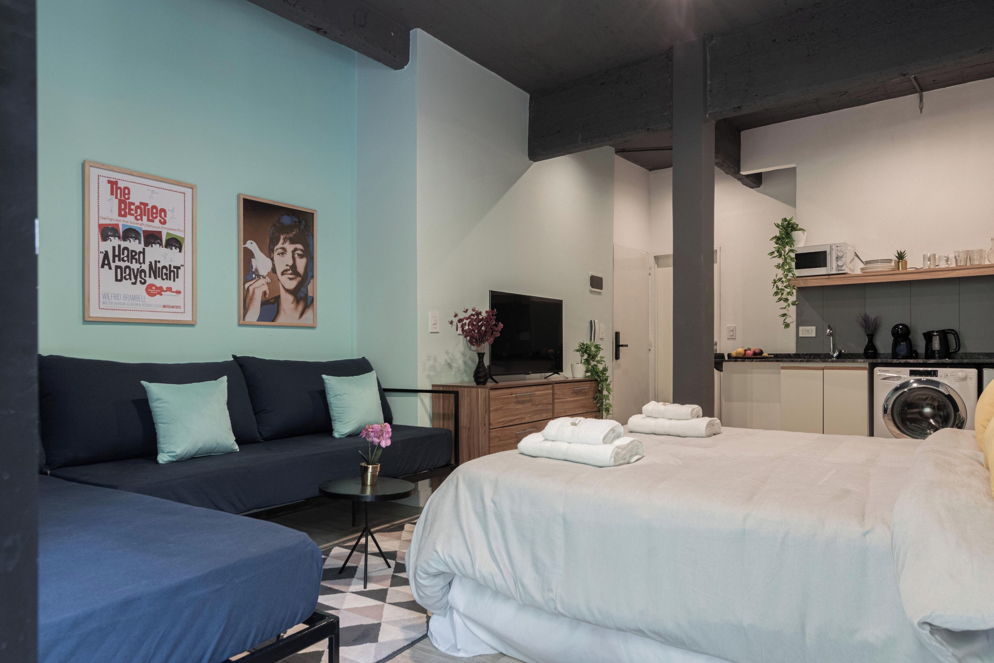 A modern hotel room with a bed, a sofa, a TV, a washing machine, and a kitchen area.
