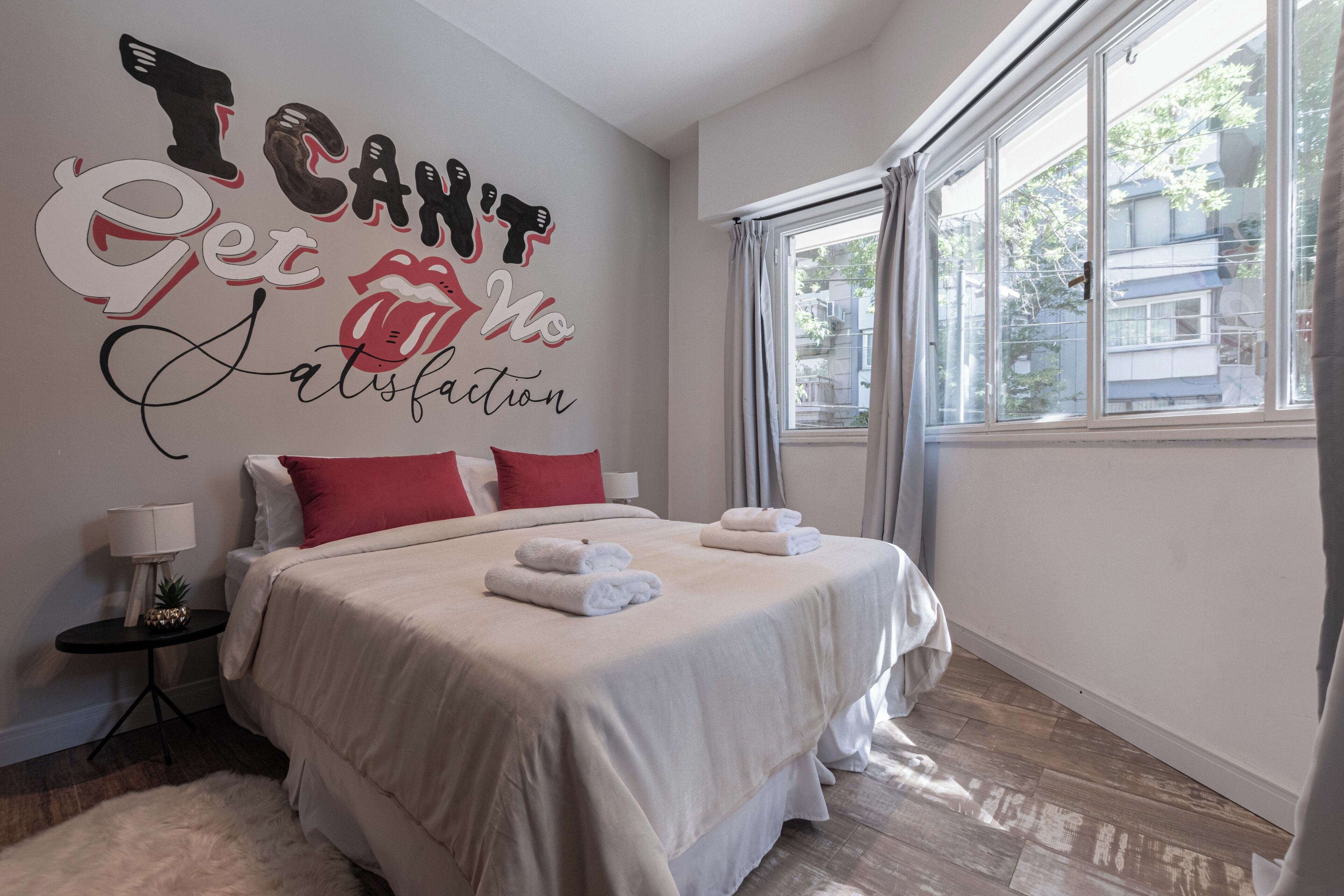 Una cama bien hecha con sábanas blancas y almohadas rojas, una pared con un texto motivacional y una ventana con cortinas.