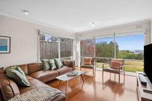 A spacious living room with a brown leather sofa, a coffee table, and a chair. There are large windows offering a view of the outdoors.