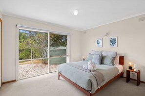 A bedroom with a large sliding glass door leading to a covered outdoor area, a bed with a wooden frame, two bedside tables with lamps, and two wall-mounted artworks. - Surfside Serenity (Anglesea)
