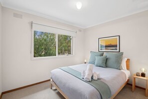 Een slaapkamer met een bed, een raam met uitzicht op bomen, een lijst met een afbeelding aan de muur en een nachtkastje met een lamp.
