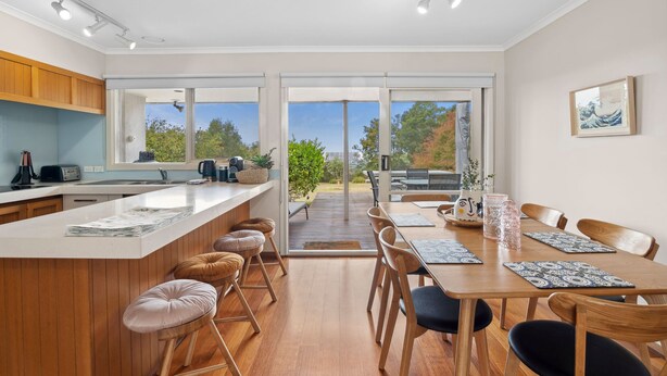 A modern kitchen with a large island, a dining area with a wooden table and chairs, and a view of the outdoors through large sliding doors. - Surfside Serenity (Anglesea)