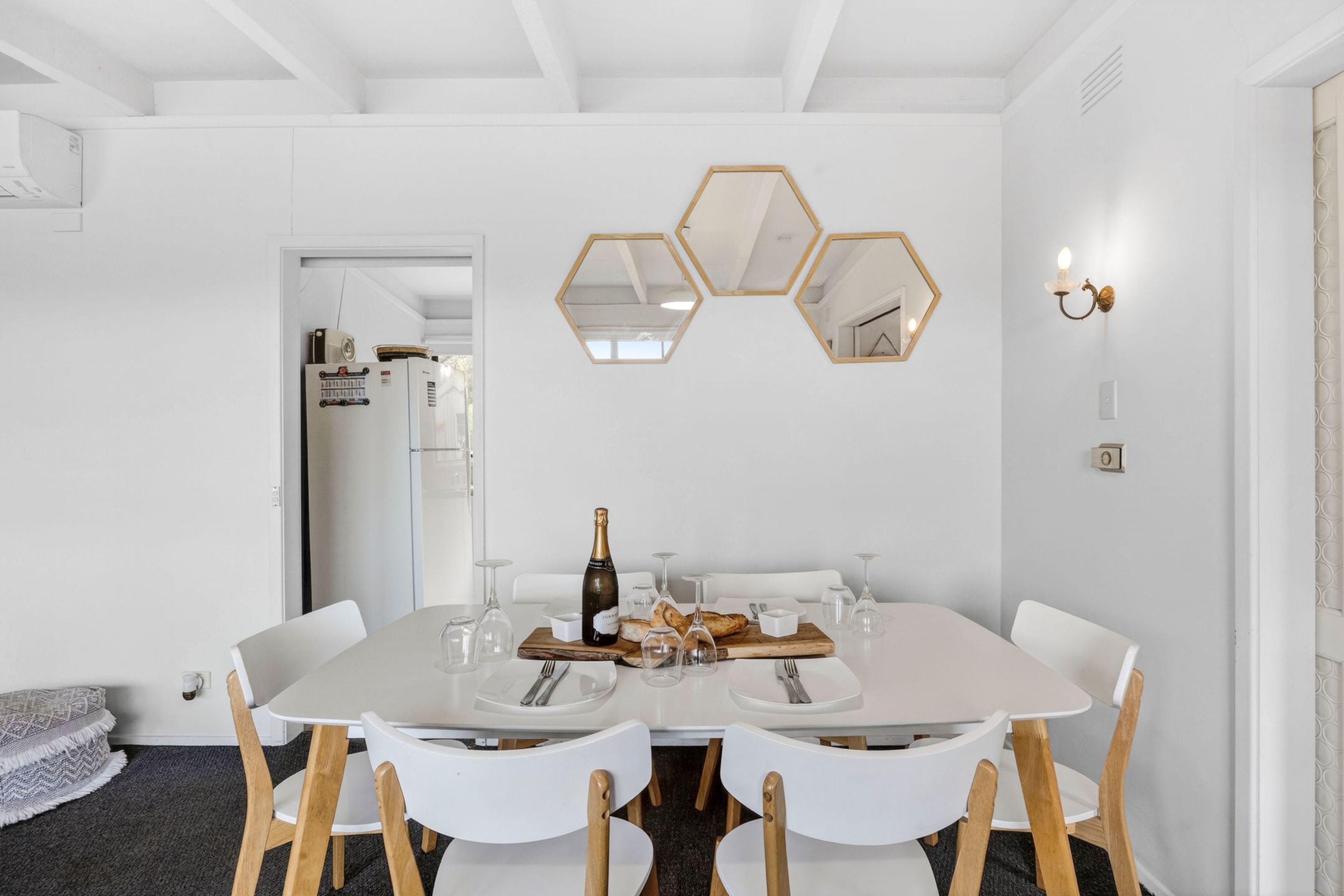 A dining area with a white table set for four, wooden chairs, and a hexagonal mirror on the wall.