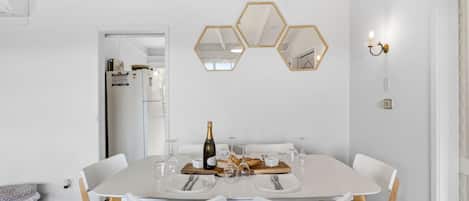 Un coin repas avec une table blanche dressée pour quatre personnes, des chaises en bois et un miroir hexagonal accroché au mur.