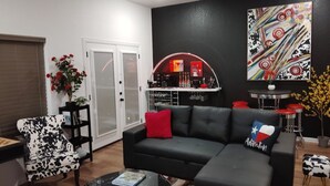 A living room with a black leather sofa, a black and white patterned armchair, a glass coffee table with books, a red cushion, and a Texas-themed pillow. - Walk to the Arlington stadiums from this Luxurious Condo w/ Premium Amenities! Rooftop Retreat w views of stadiums, private garage, gourmet kitchen & gated entry. Book Now to stay at the New Ruby Lux Skyline Retreat  in the Heart of the Arlington! (ARLINGTON)