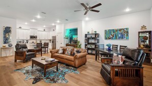 A spacious living room with a brown leather sofa, a wooden coffee table, and a leather armchair. There is a dining area with a wooden table and chairs. The kitchen is visible in the background with white cabinets and stainless steel appliances.