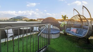 A rooftop terrace with artificial grass, a hanging chair with an American flag pillow, and a round seating area with cushions.