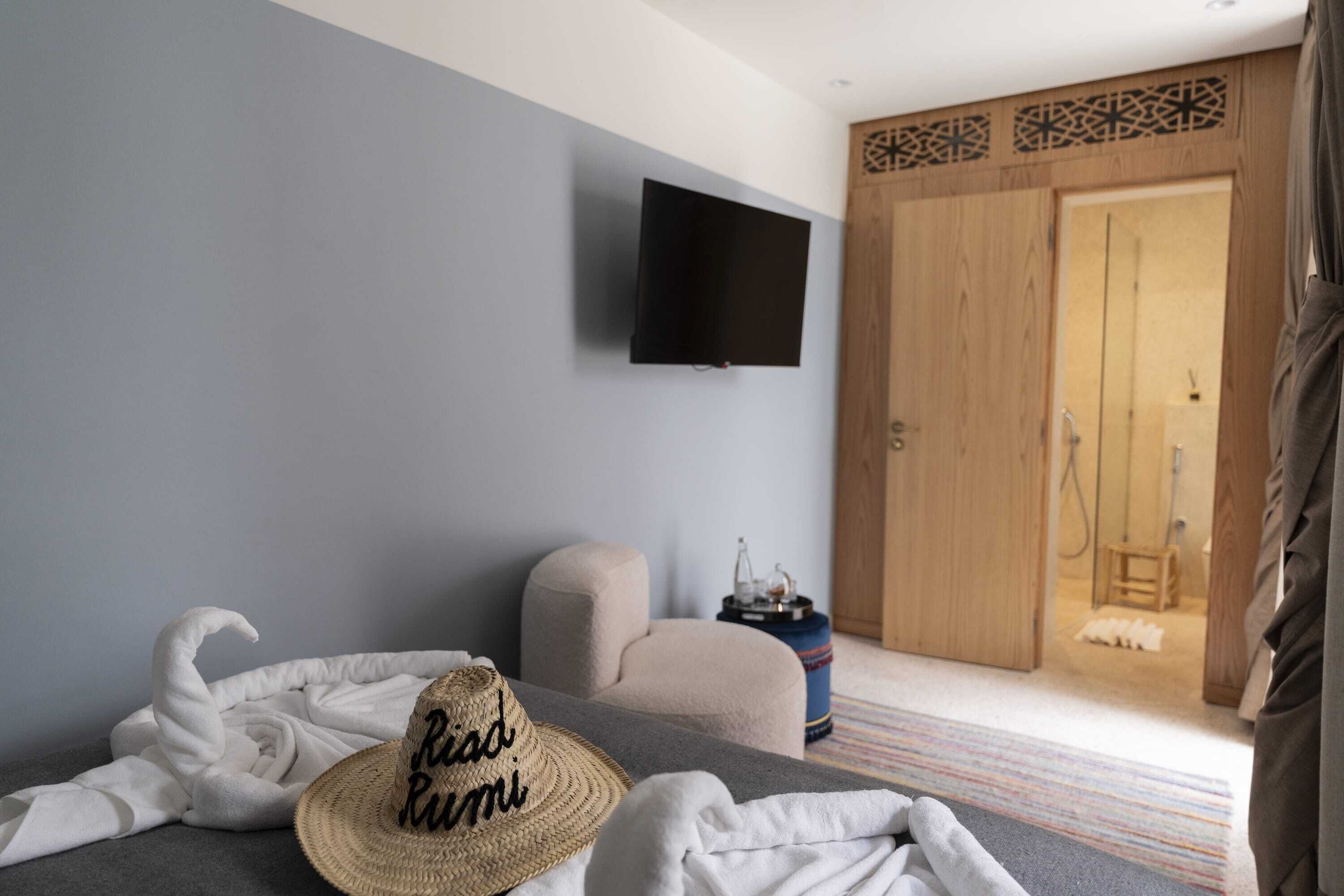 A bedroom with a bed, a television mounted on the wall, a wooden wardrobe, and a bathroom visible through an open door.