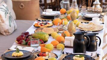 Una mesa de comedor dispuesta con frutas, bebidas y una jarrón, rodeada de sillas de mimbre y una planta en maceta.