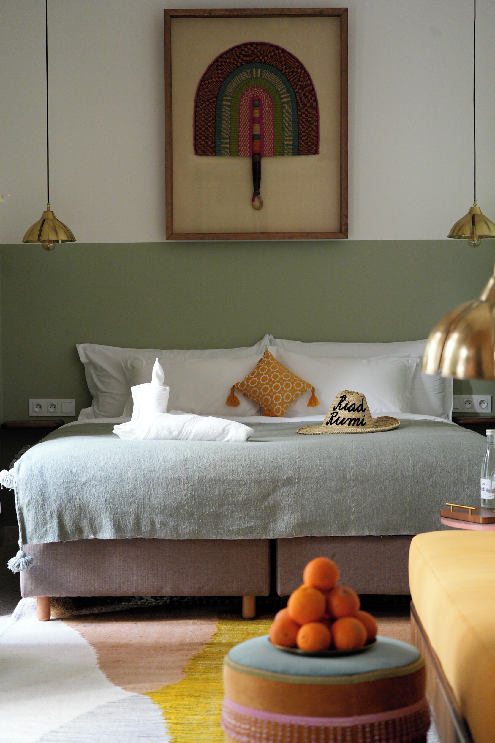 A neatly made bed with a white blanket, a towel, and a hat with the text "Road to Rome". There is a small stack of oranges on a round table beside the bed.