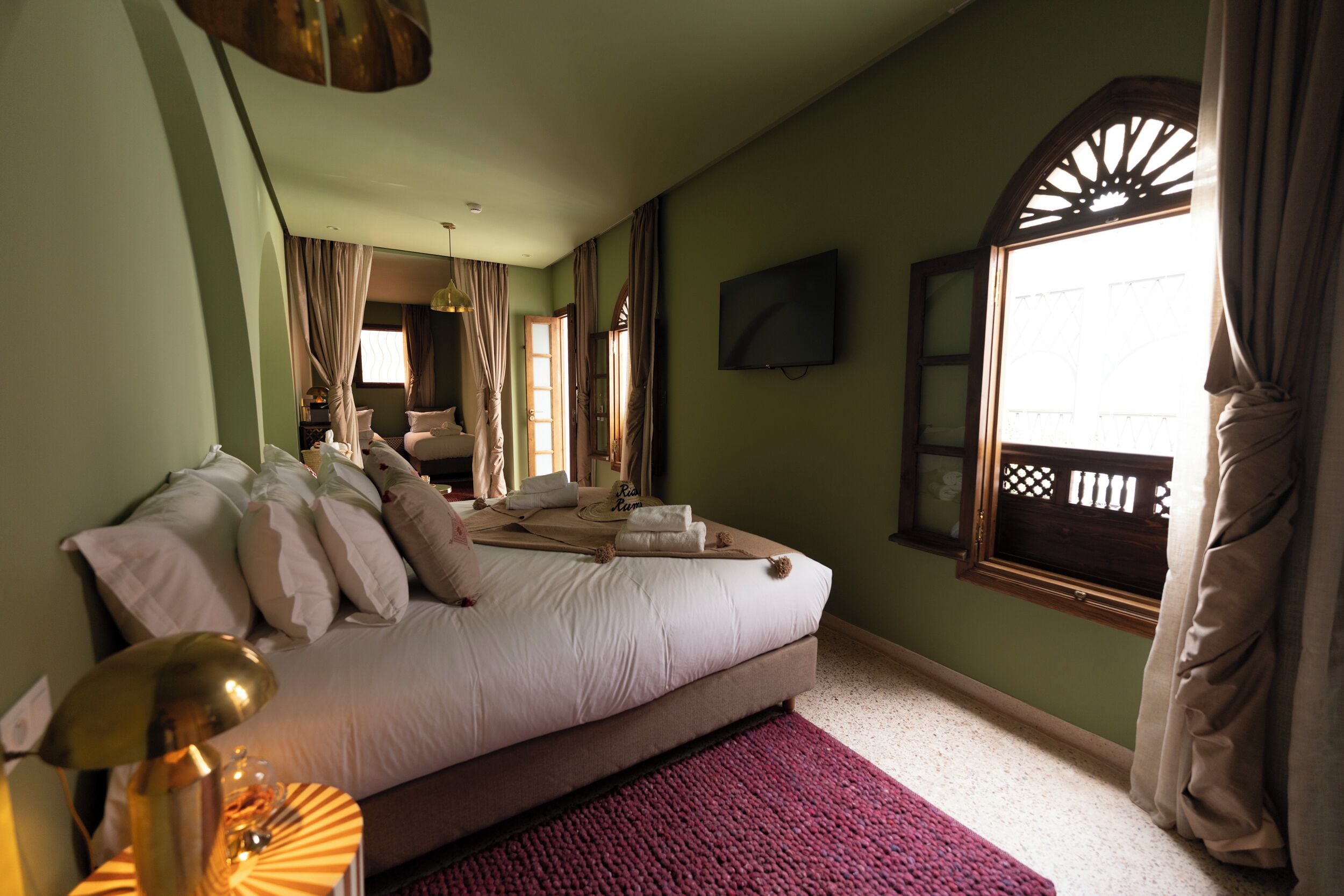 Un dormitorio con una cama grande, un televisor montado en la pared y un ventanal con cortinas.