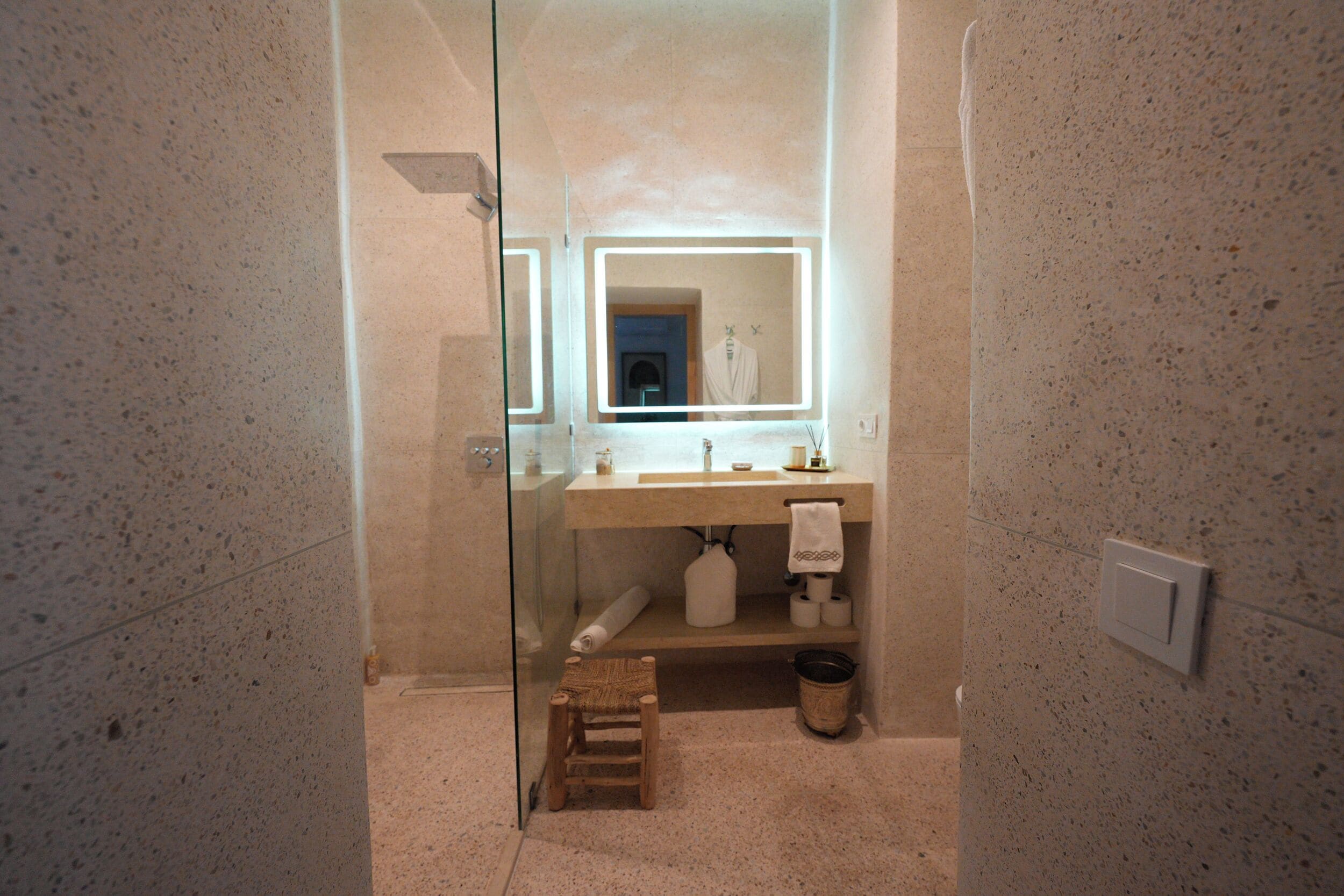Un baño moderno con cabina de ducha de vidrio, un taburete de madera, un lavabo con una toalla y un espejo con iluminación integrada.