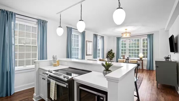 Une cuisine moderne dotée d’un îlot central, d’un four encastré et d’un micro-ondes. On y trouve des rideaux bleus, un coin repas avec une table et des chaises, ainsi qu’un téléviseur fixé au mur.