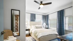 Une chambre à coucher avec un lit, un ventilateur de plafond, une table de chevet, une lampe et une fenêtre avec des rideaux.