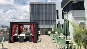 Ein Sitzbereich im Freien mit Tischen und Stühlen, ein überdachter Sitzbereich mit roten Vorhängen und ein modernes Gebäude im Hintergrund.
