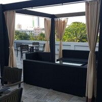 Un área de estar al aire libre con una mesa y sillas, rodeada de mobiliario de mimbre oscuro y cortinas color beige.