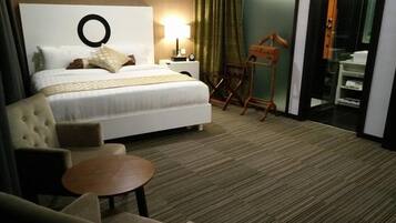 A hotel room with a bed, a round table, a chair, a lamp, a wooden chair, and a bathroom visible through a glass door.