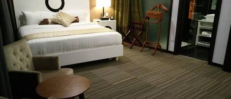 A hotel room with a bed, a round table, a chair, a lamp, a wooden chair, and a bathroom visible through a glass door.