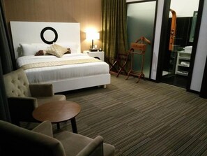 A hotel room with a bed, a round table, a chair, a lamp, a wooden chair, and a bathroom visible through a glass door.