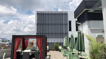Un área de estar al aire libre con mesas y sillas, un área de estar cubierta con cortinas rojas, y un edificio moderno al fondo.