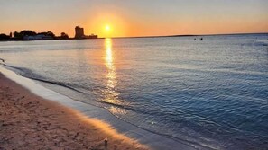A beach with a sunset, calm water, and distant buildings.
