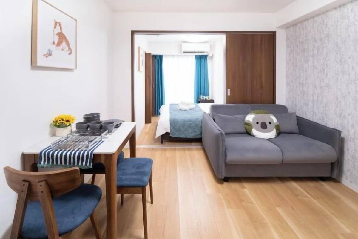 Een moderne kamer met een bank, een eettafel met stoelen, een bed met een blauwe deken en een luchtverkoeler aan de muur.