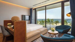 A modern hotel room with a bed, a desk, a TV, and a balcony with a view of the beach. - Beachfront Luxury Villa: Your Private Paradise (Da Nang)