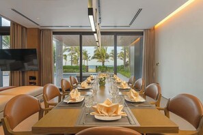 Uma sala de jantar com uma mesa comprida posta para refeição, janelas amplas com vista para palmeiras e o oceano, e uma televisão de tela plana fixada na parede.