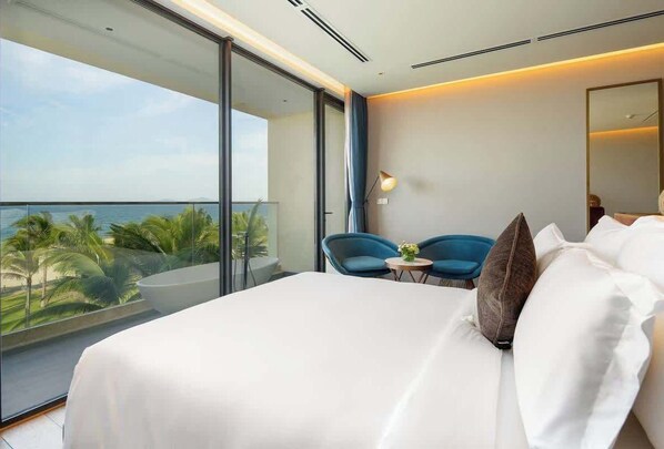 A modern hotel room with a large bed, a small table, two chairs, and a balcony with a view of palm trees and the sea.