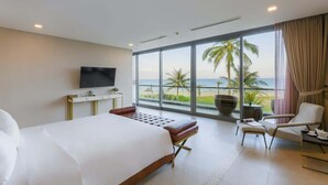 A modern hotel room with a large bed, a TV, a seating area with a view of the beach, and a balcony with palm trees. - Luxe 6BR Beachfront Villa: Group Getaway Haven (Da Nang)