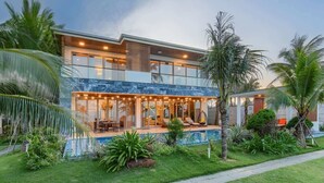 A modern two-story house with a glass facade, a swimming pool, and a well-maintained garden with palm trees. - Luxe 6BR Beachfront Villa: Group Getaway Haven (Da Nang)
