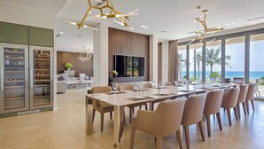 A modern dining area with a long table, multiple chairs, and a large window offering a view of the ocean. - Luxe 6BR Beachfront Villa: Group Getaway Haven (Da Nang)