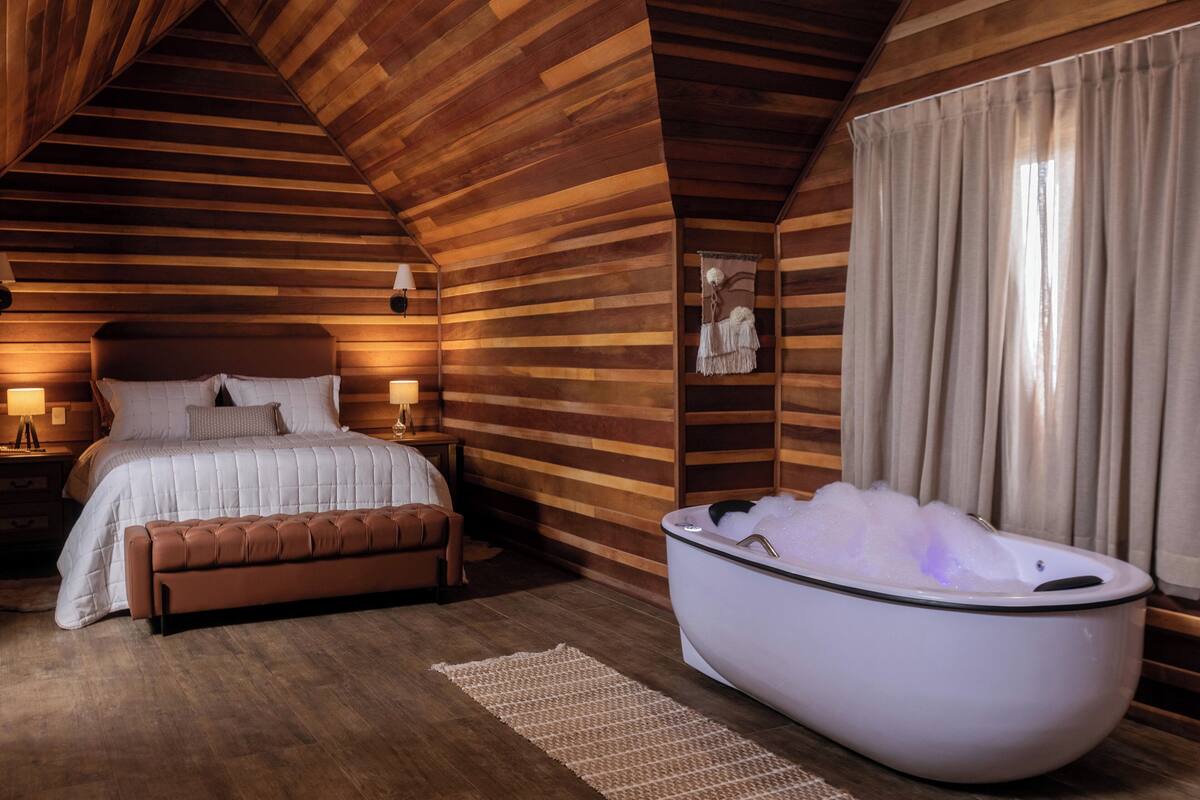 A bedroom with a wooden sloped ceiling, a large bed with white bedding, a bench, a bathtub filled with bubbles, and a nightstand with a lamp.