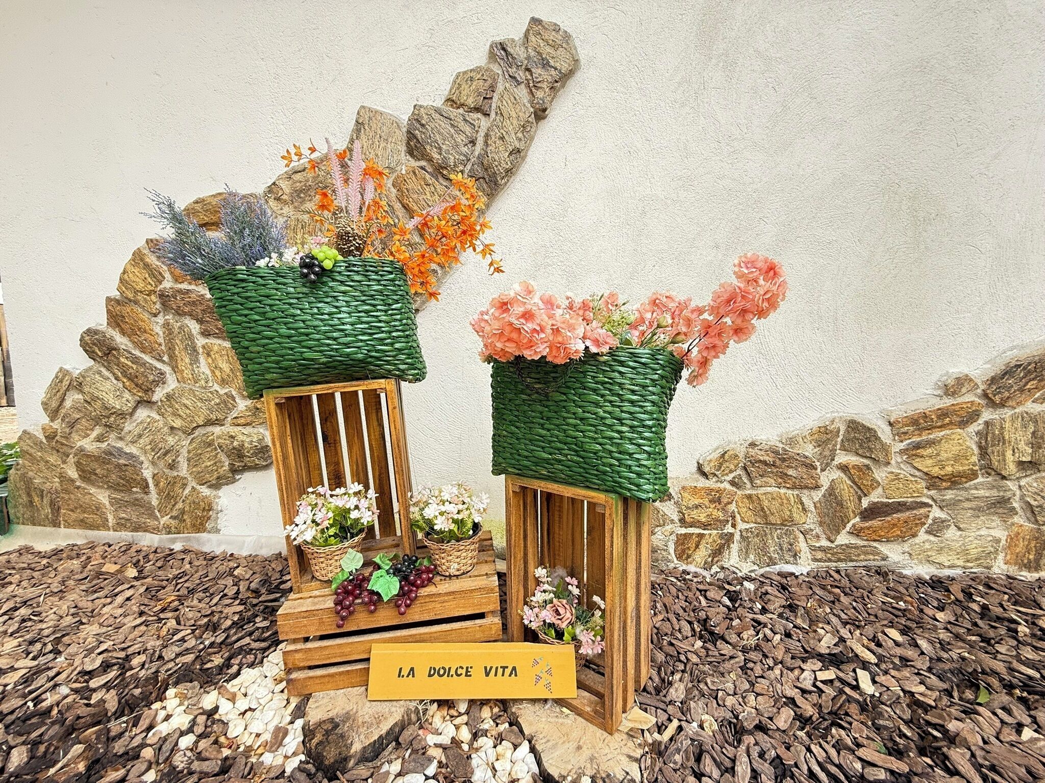 Two rustic wooden stands with woven baskets containing flowers and grapes, set against a stone wall backdrop.