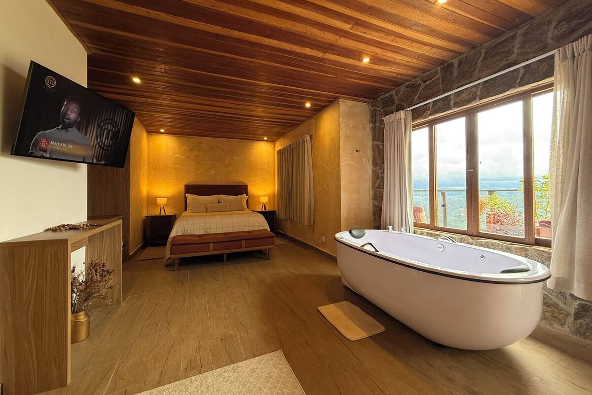 A bedroom with a large bed, a bathtub, a TV, and a view of the sea.