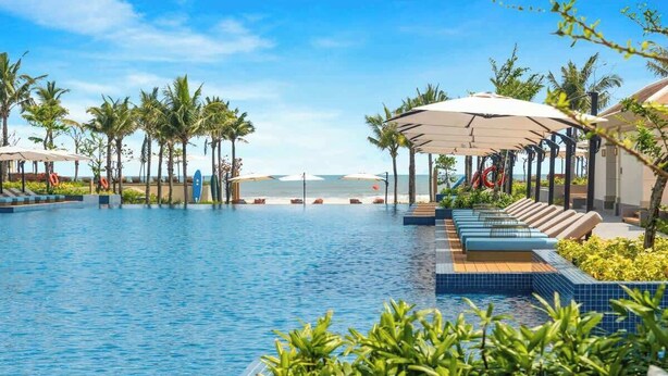 A swimming pool with lounge chairs and umbrellas, surrounded by palm trees and a view of the ocean.