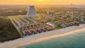An aerial view of a coastal resort with a central high-rise hotel, multiple bungalow-style accommodations, and a beachfront.