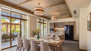 A spacious dining area with a large table set for six, surrounded by modern chairs. A kitchen with dark cabinetry and a refrigerator is visible in the background.