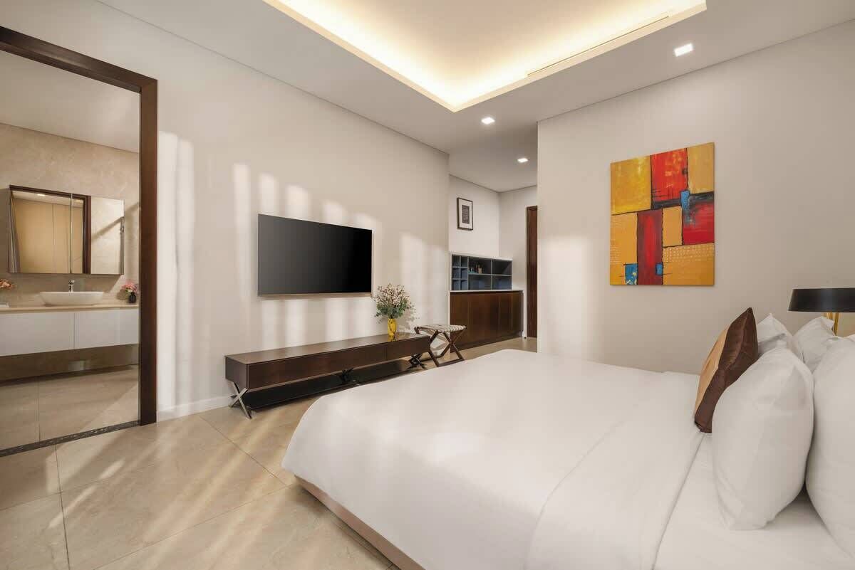 A modern hotel room with a large bed, a flat-screen TV mounted on the wall, a small seating area, and a bathroom visible through an open door.
